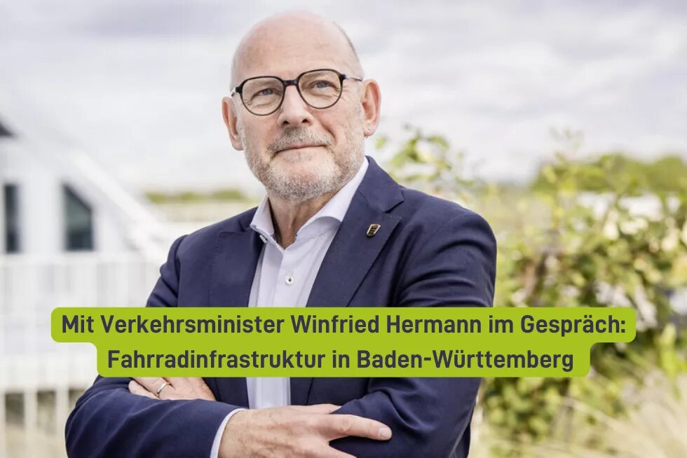 Das Bild zeigt eine Person mit Brille und einem Anstecker, die direkt in die Kamera blickt. Über der Person steht in einer Schriftleiste: "Mit Verkehrsminister Winfried Hermann im Gespräch: Fahrradinfrastruktur in Baden-Württemberg". Im Hintergrund ist unscharf eine Gebäudestruktur zu erkennen.